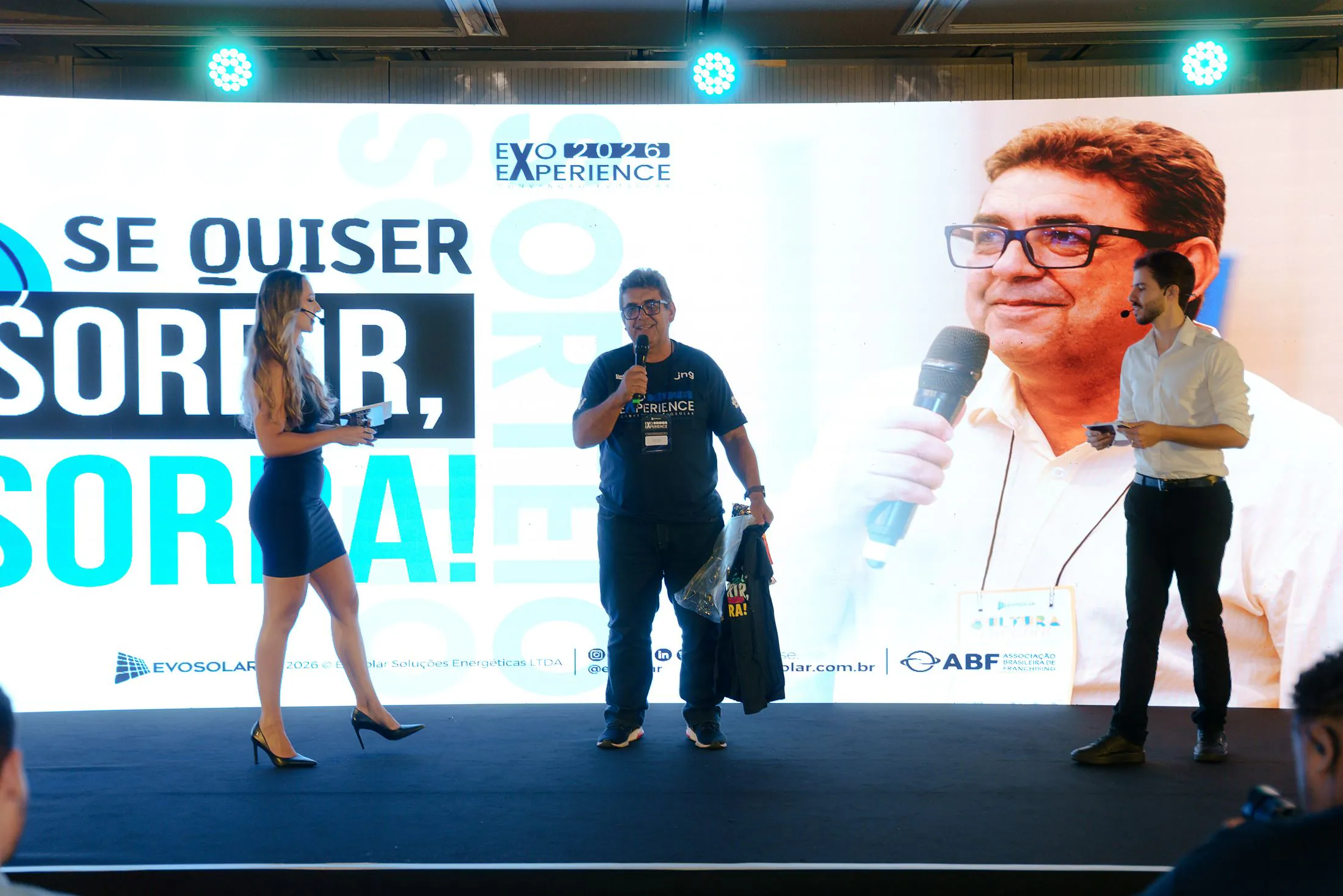 Sorteio de brindes no palco da convenção de 2026, diante do telão “Se quiser sorrir, sorra!”